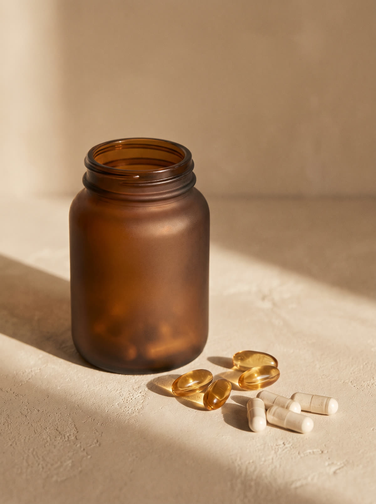 Gram Dose concept — an unbranded frosted amber glass bottle with golden softgels and bone-white capsules spilled on warm plaster in soft morning light.